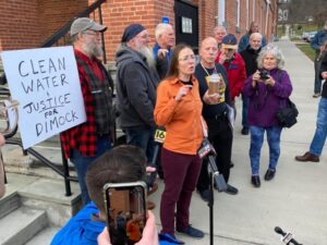 Julia Walsh speaks at microphone, supporters hold bottle of brown water and sign "Clean Water & Justice for Dimock"